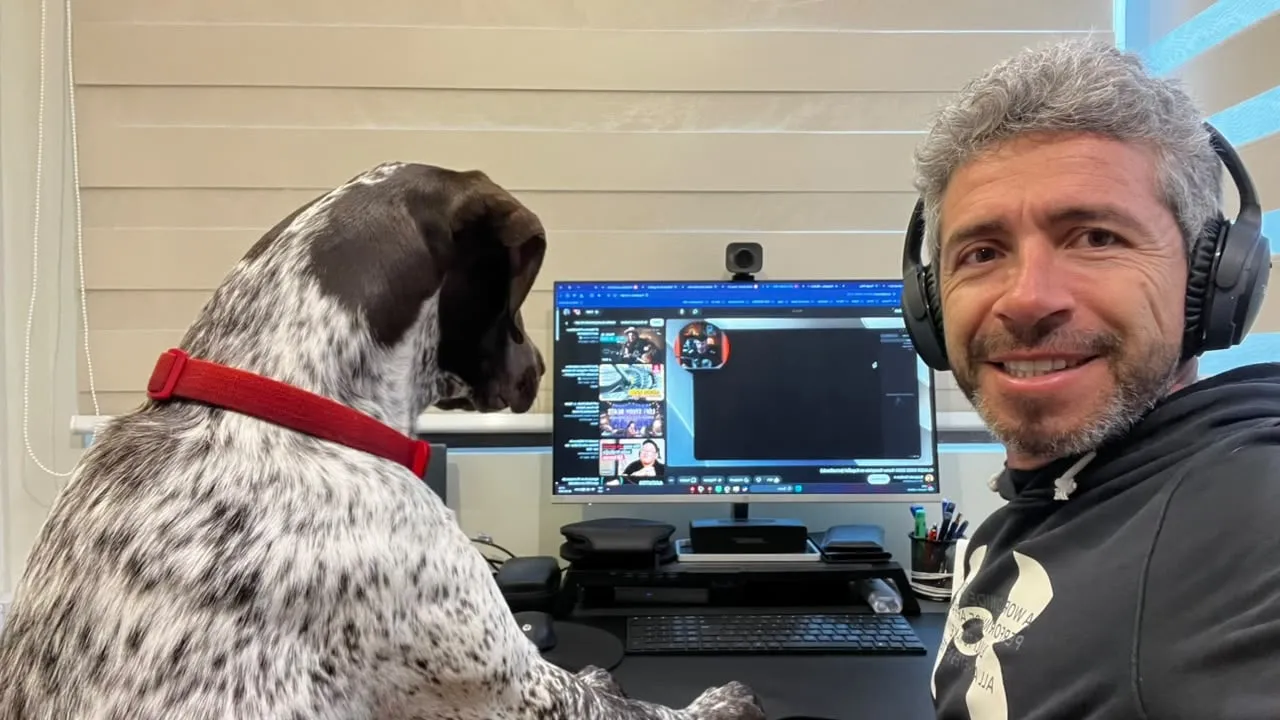 Jorge, con audífonos, sonríe a la cámara mientras Fili, con las patas sobre el escritorio, mira la pantalla del computador
